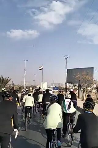 A large group of people are riding bicycles down a street. They are mostly wearing black jackets and helmets. In the distance, there's a flag and tall light posts. The sky is blue with some clouds. A drone is flying above the group. The people are cheering and yelling. There's a large billboard to the right.