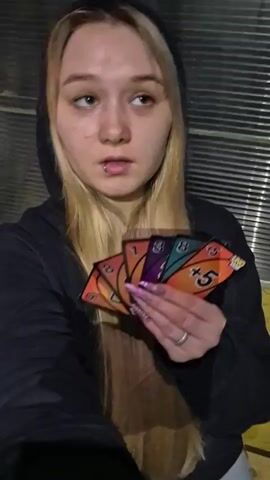 A young woman with blonde hair and a lip ring, wearing a black hooded jacket, holds a fan of UNO cards in her right hand. The cards visible are orange with a 5, purple with a 1, green with a 8, and orange with a skip symbol. Her left hand is blurred in motion. She looks at the camera with a neutral expression, then smiles slightly. The background appears to be an industrial or utilitarian setting with vertical corrugated metal siding.