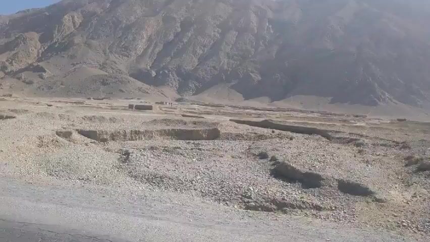 The video shows a landscape that appears to be a dry, rocky desert with a large mountain in the background. The sky is clear and blue. A person on a motorcycle drives along a dirt road in the desert. The ground is mostly covered with small rocks and some sparse vegetation. There are also some low, wall-like structures scattered across the landscape. The mountain in the background is large and rocky, with some vegetation on its slopes.