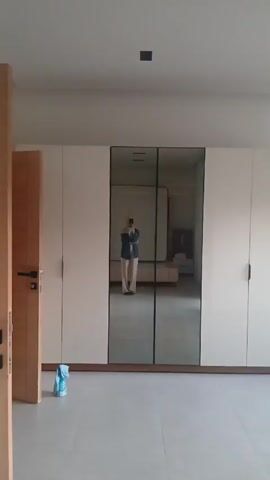 The video shows a sparsely furnished room with a large built-in closet. The closet has white doors on either side and a set of mirrored doors in the middle. A person holding a phone is reflected in the mirror, wearing a hijab, a blue shirt, and beige pants. The room is brightly lit and appears empty except for a blue package on the floor and a view into an adjacent room with a bed and bedside table visible in the mirror's reflection. The camera is static.