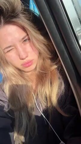 A young woman with long blonde hair is shown in a close-up shot, her face illuminated by sunlight. She is wearing headphones. She blinks and her lips are slightly parted.