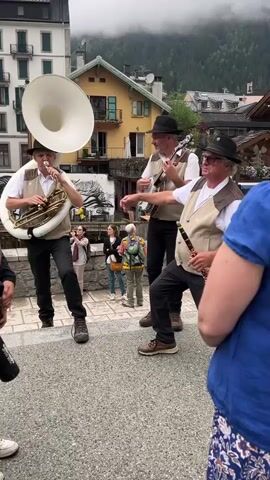 A group of three men are playing music outdoors in what appears to be a town square or street. The man on the left is playing a tuba, the middle man is playing a banjo, and the man on the right is playing a clarinet and singing. All three men are wearing hats and vests over white shirts. There are a few other people visible in the background, watching the performance. The buildings in the background suggest a European setting, possibly in the mountains. The weather appears to be overcast.