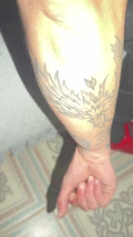 The video shows a light-skinned arm with several faded tattoos. One tattoo appears to be a phoenix-like bird, with wing and flame details. There's also a tattoo of the word "Resiliencia" written vertically, and other smaller tattoos that are difficult to decipher. The background consists of a tiled floor in neutral colors, a piece of gray fabric, and a glimpse of a red object. The hand connected to the arm is fisted, and the camera slowly moves upwards, focusing on the tattoos.