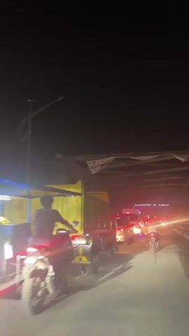 At night, the camera is positioned within a tunnel of a metal bridge, focusing on traffic moving through. A large, yellow dump truck is visible to the left, followed by a silver car and a white van equipped with blue lights underneath and red and blue flashing lights on top. Motorbikes and other cars can be seen further ahead. A banner with the words "MUATAN MEL" is hanging from the bridge's structure above. The scene is dimly lit by headlights and streetlights.