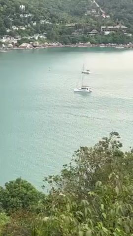 The video showcases a scenic view of a turquoise bay, captured from an elevated vantage point partially obscured by foliage. Two boats, one a catamaran and the other a sailboat, are visible in the water. The shoreline is marked by lush greenery and scattered buildings. The overall impression is one of tranquility and natural beauty.