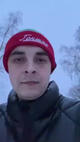A man is recording a selfie video outdoors in what appears to be a snowy or overcast environment. He is wearing a red beanie with white lettering, a dark puffer jacket, and is facing the camera. Behind him are trees without leaves and possibly snow-covered ground. He is speaking, although the audio is unclear. The focus is primarily on the man's face.