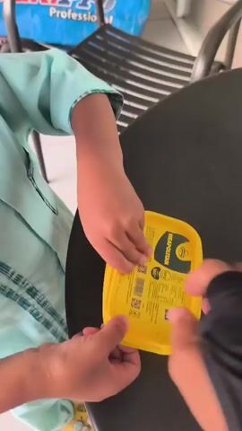 This is a close-up, overhead shot of two pairs of hands opening an ice cream container. The person on the left is wearing a light blue shirt with a textured pattern. The person on the right is wearing a black sleeve, possibly an arm covering. Both individuals are focused on the yellow-lidded container, which is placed on a dark, round table. The ice cream inside is tri-colored, with layers of pink, yellow, and brown. One lid has been fully removed and rests near the container, while the other is in the process of being opened. A wooden spoon lies on the table to the right of the container. The environment appears to be indoors, with a glimpse of a metal chair and a blue object in the background. The camera angle is steady, offering a clear view of the action.