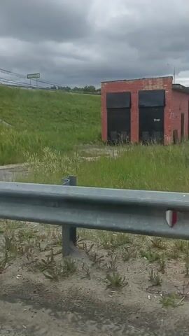 The video captures a dynamic scene from a moving vehicle, likely a car, looking towards the right side of the road. The footage opens to reveal a guardrail in the foreground, its metal structure weathered and showing signs of age. Beyond the guardrail, a patch of dirt and debris lines the roadside, hinting at an undeveloped or perhaps neglected area.

Further back, a vibrant green field stretches out, its tall grasses swaying gently, suggesting a breezy atmosphere. In the distance, atop a grassy incline, a sign with green lettering is visible, though its text is mostly obscured by the distance and angle. A red brick building with dark, boarded-up windows stands out against the natural backdrop, appearing abandoned or disused. Power lines crisscross the sky, indicating nearby infrastructure.

As the vehicle continues to move, the view shifts. The grassy incline reveals a paved pathway with concrete tiles leading up, accompanied by a set of metal railings and stairs. The sky overhead is overcast, with heavy, gray clouds dominating the frame, indicating a gloomy or possibly rainy weather condition. The time of day is unclear due to the cloud cover, but the lighting suggests it is daytime. The presence of a guardrail and a road implies this is a roadside view, possibly on the outskirts of a town or in a rural area. There are no discernible people or animals in the frame, and the overall mood is somewhat desolate due to the abandoned building and overcast sky.