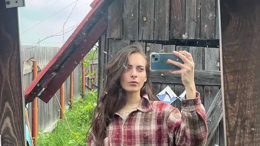 A young woman with long, dark brown hair is taking a selfie in a mirror. She is wearing a plaid shirt in shades of red, brown, and tan. The background appears to be an outdoor setting with a rustic wooden fence and some greenery. The woman is holding a smartphone in her right hand, which is angled to capture her reflection. She is looking at the camera and has one hand on her hip. The mirror is framed by rough-hewn wood. There are some metal fixtures visible to the left of the mirror. The lighting suggests it is daytime.