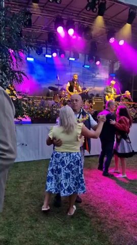 The video shows an outdoor event with a live band performing on a stage. In the foreground, a couple is dancing closely on a grassy area. The woman is wearing a yellow top and a blue and white patterned skirt, with her hair blonde. The man is wearing a white t-shirt, a navy vest, and plaid shorts, with a lei around his neck. Another couple is dancing in the background to the right, with the woman in a red jacket and a black and white skirt, and the man in a dark shirt. The stage is lit with colorful purple and blue lights, and the band members, including guitarists and a drummer, are visible. There are also plants and flowers decorating the stage. The overall atmosphere appears festive and social.