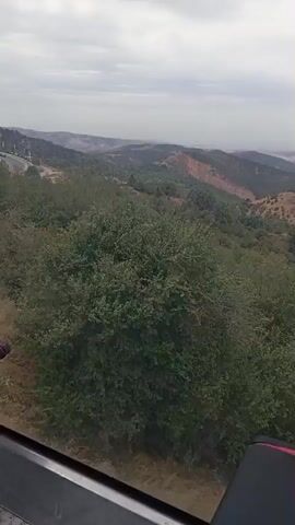 The video shows a view from a cable car moving through a mountainous, arid landscape. The cable car has dark seats with red accents. In the foreground, lush green bushes and dry grasses are visible. In the background, rolling hills and mountains with patches of greenery and barren earth stretch into the distance under a cloudy sky. Another cable car can be seen in the middle ground, moving along a cable. A building with a green roof and a curved road are also visible on the hillside.