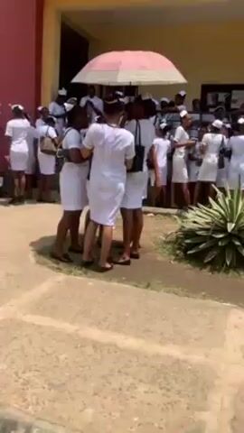 A group of people dressed in white uniforms, likely nurses or medical personnel, are gathered in front of a building with a red and beige exterior. They are standing in a small group, some with handbags, on a paved area. An umbrella provides shade. The camera moves along a walkway before focusing on a large group of people in similar white uniforms gathered on a grassy area under trees. A person in a red shirt and black shorts walks in front of the group.