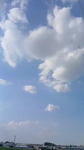 This media file captures a serene daytime scene under a bright blue sky adorned with fluffy white clouds. The camera pans slightly, revealing a wide expanse of sky with varying cloud formations, from large, cumulus clouds to smaller, scattered wisps. 

The lower portion of the frame shows a landscape that appears to be a rural or semi-urban area in Daliguzar, Uzbekistan. A green field, possibly of crops, occupies the foreground, suggesting a natural or agricultural setting. Behind the field, a line of buildings and structures is visible, including what look like industrial buildings or warehouses, along with some smaller houses and a tall telecommunications tower or antenna. Trees are interspersed among the buildings.

The time of day is clearly daytime, likely late morning or afternoon, given the bright sunlight and the clear visibility. The weather appears to be pleasant, with a clear sky and scattered clouds, indicative of a fair-weather day. The overall mood is calm and tranquil, evoked by the vastness of the sky and the quiet landscape. There are no people visible in the frame, and no discernible text is present. The focus is on the natural beauty of the sky and the subtle presence of human development in the background.