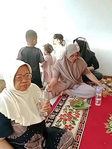 A group of people are sitting on a red carpet with a floral pattern, eating. The carpet is adorned with a large oval floral design in the center. In the foreground on the left, an elderly woman wearing glasses and a light-colored headscarf holds a glass of water. Next to her, a woman in a beige hijab and matching outfit is seated, looking down at food on a banana leaf. To her right, a woman in a black hijab is also seated. Further back, two children are standing. One child in a dark shirt stands facing away from the camera, while a younger child in a light-colored outfit stands next to them. A man in a gray t-shirt and white cap is seated in the middle ground, eating with a utensil. Another person, partially visible on the right, is holding a banana leaf with food. Several bottles of water and plates with food are scattered on the carpet. The environment appears to be indoors, with a plain white wall in the background.
