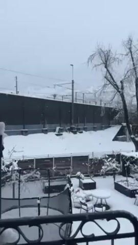 The video shows a snow-covered backyard scene, filmed from a balcony. Snow is actively falling. In the foreground, a metal balcony railing is visible. The backyard contains a trampoline, a small round table with chairs, and some potted plants, all covered in snow. A wooden fence separates the backyard from a slightly elevated area covered in snow. Behind the elevated area, there is a tall, dark, noise barrier and some utility poles with wires. A bare tree is visible to the right. The sky is overcast and grey.
