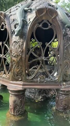 A bronze fountain structure is partially submerged in green water. The structure is ornate, featuring intricate scrollwork, stylized leaves, and a domed roof adorned with what appear to be bronze lizards. Within the open framework of the fountain's central section, a small, dark figure is visible. The figure appears to be seated and bent over, possibly working or interacting with something within the structure. Water gently spills from the fountain into the surrounding pool. The overall setting suggests an outdoor environment, possibly a garden or park, with greenery visible in the background.