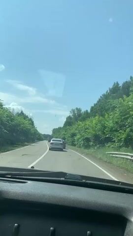 The video is a first-person perspective from inside a car driving on a paved road surrounded by lush green trees. Several cars are visible ahead on the road, including a white sedan and a red car. The sky is clear with scattered white clouds. The video captures the motion of the car as it travels along the scenic route.