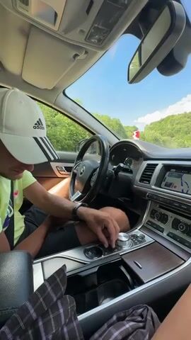 The video shows the interior of a car, viewed from the passenger seat. A person wearing a white Adidas baseball cap and a green shirt is in the driver's seat, operating the gear shift. The car is moving, as indicated by the scenery outside the windshield and side windows, which includes trees, a blue sky with clouds, and a gas station with red and white awnings. A motorcycle rider is visible on the right side of the frame, stopped at what appears to be a gate. The car's dashboard and navigation screen are also visible. The overall environment suggests a sunny day and travel through a rural or semi-rural area.