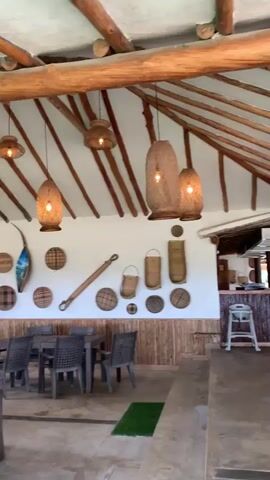 The video pans across an open-air dining area with a thatched roof supported by wooden beams. Several tables and chairs are visible, arranged under the canopy. In the background, a counter or bar area is set up, with a high chair nearby. The walls are decorated with woven baskets and other rustic items. The scene transitions to show more dining tables and chairs, some under thatched roofs, set amidst lush greenery and palm trees. Further into the video, the view opens up to a sandy beach area with more palm trees and people walking in the distance. The overall environment appears to be a resort or a beachfront restaurant.