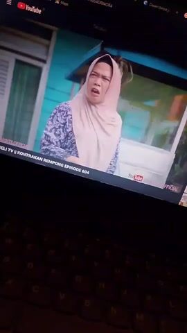 A person wearing a headscarf and a patterned shirt is looking at the camera with a surprised expression. The person has a short hairstyle. The background appears to be outdoors, with a building and some plants visible. The video is being viewed on a laptop, with part of the keyboard visible in the foreground. The YouTube interface is visible on the screen, indicating that the video is being played from YouTube. The title of the video appears to be "KONTRAKAN REMPONG EPISODE 604". The timestamp indicates the video is at the very beginning.