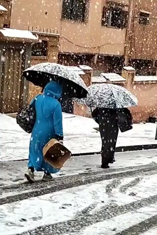 The video shows a snowy street scene with two people walking under umbrellas. Snow is falling heavily. The person on the left is wearing a blue robe and carrying a patterned tote bag on their shoulder and a tan box in their other hand. The person on the right is wearing a black robe. Both are walking away from the camera and across a street covered in snow, leaving tire tracks in the snow. Buildings line the street, and snow is accumulating on their roofs and ledges. A car is parked along the street in the distance. A utility pole is visible.