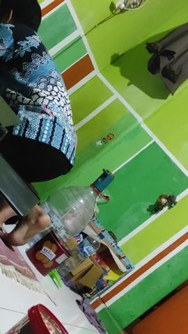 Here is a description of the video.

The video depicts a man dressed in a blue batik shirt and black pants standing in a room with green walls and a white tiled floor. The walls are painted with a design of horizontal stripes. Behind him are a few objects, including a large plastic water bottle, boxes, and a red cookie tin labeled "REGAL." A jacket hangs on the wall. The camera angle is slightly low, and it pans up to follow the man as he straightens his back and turns to look towards the left side of the frame.