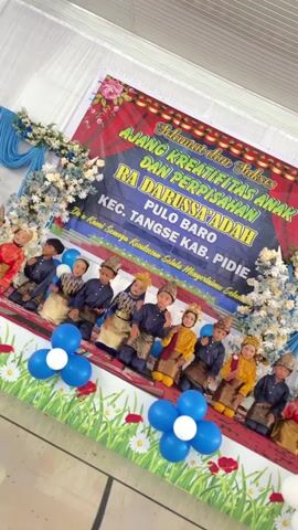 A group of children, mostly boys, are sitting on a stage in what appears to be an indoor event. The children are dressed in traditional attire, some with head coverings. Behind them, a large banner reads "SELAMAT DAN SUKSES AJANG KREATIFITAS ANAK DAN PERPISAHAN RA DARUSSA'ADAH KEC. TANSE KAB. PIDIE". The stage is decorated with white and blue flowers and balloons. The lighting is bright, suggesting artificial illumination. The overall scene depicts a cultural or educational event for young children.
