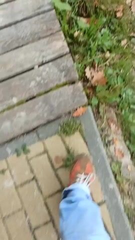 The video shows a first-person perspective of someone walking on a wooden path and then onto a dirt path. The person is wearing blue jeans and orange sneakers. The environment appears to be outdoors, with grass and fallen leaves visible on the ground. The wooden path is made of planks. The dirt path is uneven and shows footprints.