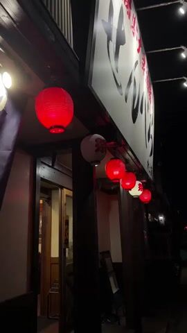 The video shows the exterior of a building at night, likely a restaurant or shop, with a large white sign featuring Japanese characters in black and red. Several red paper lanterns are hanging from the awning above the entrance, and one white lantern with red characters is also visible. The entrance appears to be a wooden doorway. The scene is dimly lit, with some string lights visible in the upper right corner. The overall impression is that of a traditional Japanese establishment at night.