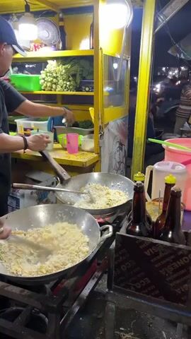 A street vendor is cooking in a food cart. The cart is yellow and illuminated by lights. In the foreground, two woks are on a stove, filled with rice. The vendor, wearing a black shirt and a white cap, is stirring the rice in the left wok with a large spatula. Another person is visible in the background, preparing ingredients. There are bottles of sauce and various containers on a shelf behind the woks. The scene appears to be at night, with a street and passing vehicles visible in the background.