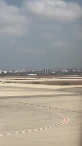 The video shows an aerial view of an airport runway. An airplane is visible in the distance, on the runway. The sky is partly cloudy, and a city skyline is visible in the background. The focus is on the runway and the airplane, with the city and sky serving as a backdrop. The scene is calm and still, suggesting a routine moment at the airport. The lighting suggests it is daytime, possibly late morning or early afternoon.