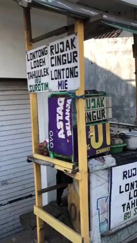 A street food stall with a yellow wooden frame is visible. Two signs are attached to the frame. The left sign lists "LONTONG RUJAK LODEN TAHULEK IBU SUROTIN". The right sign lists "CINGUR LONTONG MIE". Below the signs, a purple and green box advertising "ASTAGA BELANJA" is attached to the stall. A person, partially visible, stands behind the stall. Another sign, partially obscured, is attached to the lower front of the stall and displays "RUJAK LONTONG TAHU BU SURO". The stall is positioned in front of a closed metal roller door.