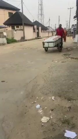 A man in a red shirt and blue jeans pulls a two-wheeled cart loaded with white jerrycans down a street in a residential area. Another person walks in the distance. Buildings are visible on either side of the street. A tall utility tower stands in the background.