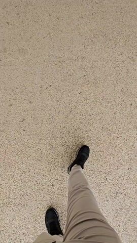 The video starts with a view of a person's feet standing on a speckled, light-colored concrete surface. The person is wearing khaki-colored pants and black sneakers. The camera slowly pans up to reveal an outdoor corridor between two rows of white buildings with shops or residences. The buildings have a Mediterranean architectural style with arches and window awnings. The corridor is paved with the same speckled concrete and has manicured hedges and trees lining the walkway. In the distance, there is a building with a clock tower that appears to be the focal point of the area. The sky is overcast. The camera continues to pan up and forward, further revealing the buildings and landscaping.