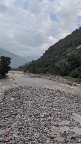 The video shows a view of a rocky riverbed with a cloudy sky above. A fast-flowing river with murky brown water moves through the scene. Lush green trees and vegetation cover a steep mountainside that rises on the right side of the river. The terrain suggests a natural, mountainous environment. There are power lines visible in the upper portion of the frame, indicating some human infrastructure in the area. No people are visible in the video. The camera appears to be stationary, capturing a panoramic view of the landscape.
