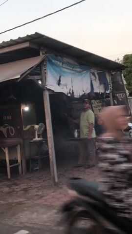 The video shows a street-level view of a small outdoor food stall or shop. In the foreground, a person wearing a light green t-shirt and dark pants stands near the stall, holding a bottle. The stall has a corrugated metal roof and appears to be constructed from various materials, including wood and possibly corrugated metal for siding. There are signs with text in what appears to be Indonesian, a fan, and some items displayed on shelves. The ground in front of the stall is paved with brick-like material and appears wet or dirty. In the background, across a grassy field, several people are visible, some sitting on benches and others appearing to be playing or observing an activity. A motorcycle is parked near the benches. The sky is overcast. The video has a slightly shaky quality, suggesting it was recorded handheld.