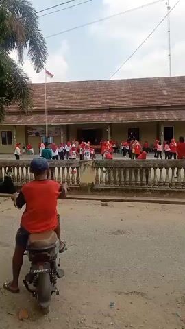 A group of people, mostly women and girls wearing red shirts and headscarves, are participating in an outdoor activity in front of a building with a red tiled roof and a white flag pole flying a red and white flag. Some of the participants are holding up numbers. A young person wearing a red shirt and a blue cap is sitting on a motorbike in the foreground, facing away from the building and looking towards the group. The ground in front of the building is paved, while the area in the foreground appears to be unpaved and dusty. The sky is clear and bright, suggesting it is daytime.