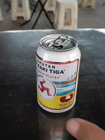 The video shows a close-up of a person's hand holding a can of "Larutan Cap Kaki Tiga" on a dark surface. The can is white with red and blue branding, featuring text in what appears to be Malay or Indonesian and an image of three running figures. The can is open, and the person's fingers are visible around its base. In the background, blurred out, are white plastic chairs and table legs, suggesting an outdoor or casual dining setting. The camera angle is slightly downward, focusing on the can and the hand.