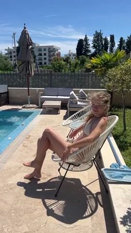 A young woman sits in a white, woven chair next to a swimming pool on a sunny day. She is wearing a light-colored bikini and has her legs crossed. Her hair is blowing in the wind. In the background, there is a pool, lounge chairs, an umbrella, and some buildings. A blue towel is on the ground next to her chair. The scene appears to be a resort or a private residence.