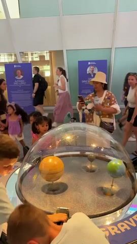 The video shows an interactive exhibit in what appears to be a science museum. In the foreground, a large, clear dome covers a circular display with a granular surface. Inside the dome, two spheres are visible on stands: a larger, yellowish-brown sphere to the left, resembling a planet with rings, and a smaller, greenish-blue sphere to the right, representing Earth. The granular surface has indentations, suggesting the exhibit demonstrates gravitational pull. Several people are in the background, some of whom appear to be looking at the exhibit. A woman wearing a hat and a patterned shirt is holding a phone and taking a picture or video of the exhibit. A young girl in a pink shirt is also observing the exhibit closely. In the background, there are informational posters with images and text. The environment seems to be well-lit and public.