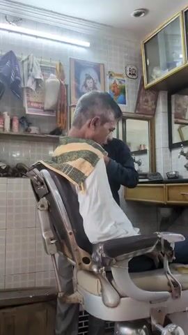 A man sits in a barber chair covered with a towel. He appears to be touching his forehead. Another person, partially visible, stands behind him and seems to be touching the first man's head. The barber shop has tiled walls and a shelf with various items. Pictures hang on the wall.