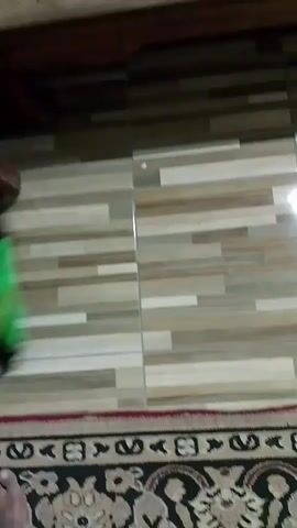 A blurry video shows a green broom being used to sweep a tiled floor. The broom is moving rapidly from left to right across the frame. A portion of a rug is visible at the bottom of the frame. The flooring appears to be a light-colored tile with a wood-grain pattern. The upper part of the video shows a dark-colored surface, possibly a piece of furniture. The video is extremely short, lasting only a few seconds.
