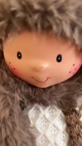 The video shows a close-up, then a wider shot, of a doll dressed in winter clothing. The doll has a light-colored face with rosy cheeks, black eyes, and a smiling mouth. It is wearing a white knitted hat with a brown fur trim, a brown fur scarf, a white knitted sweater with a diamond pattern, and brown knitted overalls with two wooden buttons. The doll also has what appears to be a brown object with a pink tip held in its right hand. The doll is positioned on a light brown, possibly leather, surface.