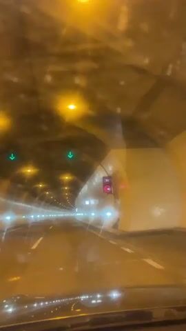 The video shows the interior of a tunnel as seen from a moving vehicle. The tunnel has a curved ceiling with evenly spaced, bright yellow lights. The walls are a mix of gray and yellow tones. There is a single white line dividing the road. Traffic signs and speed limit indicators are visible on the right wall of the tunnel. A green arrow sign hangs from the ceiling near the start of the tunnel. The vehicle appears to be moving at a moderate speed. There are water droplets on the windshield, possibly due to rain or humidity.