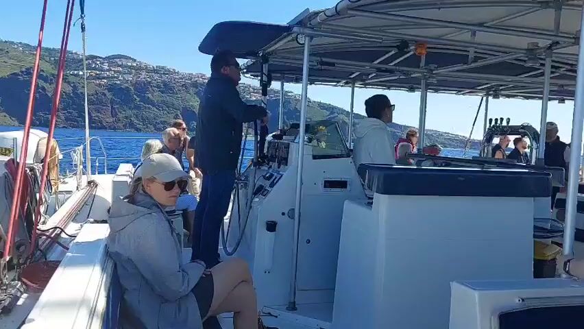 The video shows the interior of a boat filled with passengers. A man is steering the boat. Passengers are seated on benches on either side of the boat, looking out at the scenery. Some passengers are talking to each other. The boat has a canopy structure overhead, supported by metal beams. The boat is moving, and the scenery outside includes water and a distant hillside with buildings. The sky is clear and blue.