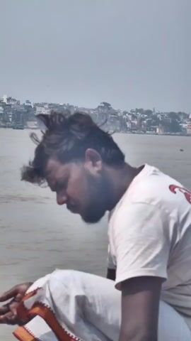 A man in a white t-shirt and white pants is sitting on the edge of a wooden boat. His head is bowed, and he appears to be looking at something in his hands. The boat is on a body of water, possibly a river, with a city visible in the background. The sky is blue with some clouds. Another person's head is visible in the foreground.