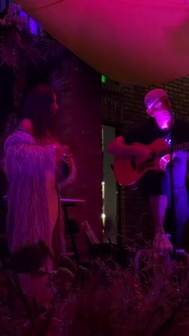 A woman with long dark hair wearing a fluffy pink robe and a light-colored dress is standing to the left, holding a microphone to her mouth. She is facing a man seated on a stool to the right, playing an acoustic guitar. The man is wearing a black t-shirt, dark shorts, a white beanie, and white sneakers. He is looking towards the woman. The scene is illuminated by purple and pink lights. A brick wall is visible behind the woman, and foliage is in the foreground. A yellow fabric structure is overhead. The overall impression is of an outdoor performance setting at night.