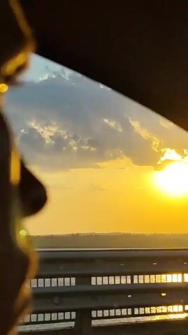 From the perspective of a moving vehicle, a vivid sunset dominates the frame. The large, bright sun glows intensely through the clouds, casting a golden hue across the horizon. A metal guardrail runs along the roadside, reflecting shimmering light from the water below, which is possibly a lake or a river. The sky is adorned with patches of clouds partially obscuring the sun. A car's side mirror is visible in the right of the frame.