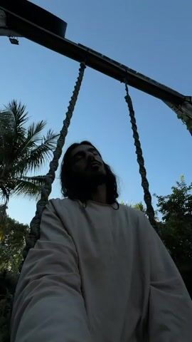 A low-angle, medium shot shows a man with long, dark hair and a beard, wearing a light-colored robe, sitting on a swing. The swing is suspended from a wooden beam by two thick ropes. The man's eyes are closed, and his head is tilted back. The background consists of a clear blue sky, lush green foliage, and part of a tiled roof. The lighting suggests it is either early morning or late afternoon. The sign above the swing is partially visible with text that is difficult to read.