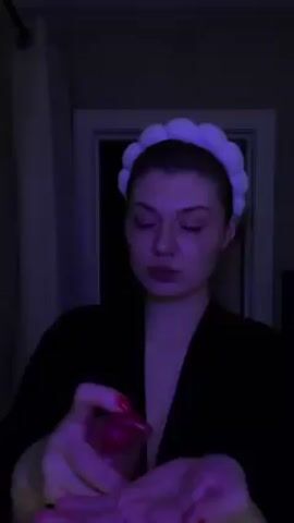 A woman with a white headband on her head is applying a liquid from a pump bottle to her face. She is wearing a black robe and standing in a dimly lit bathroom with a white door behind her. The light in the room is a dark purple. She rubs the liquid into her face, looking down as she does so.